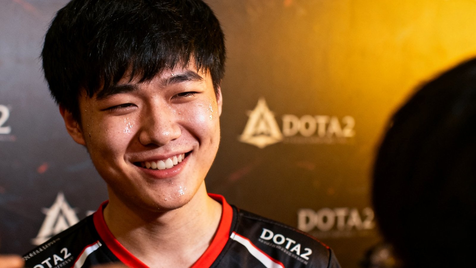 DOTA2国际邀请赛淘汰赛阶段，中国战队选手在赛后采访区面对镜头露出疲惫但满足的微笑，背景是赛事官方背景板
